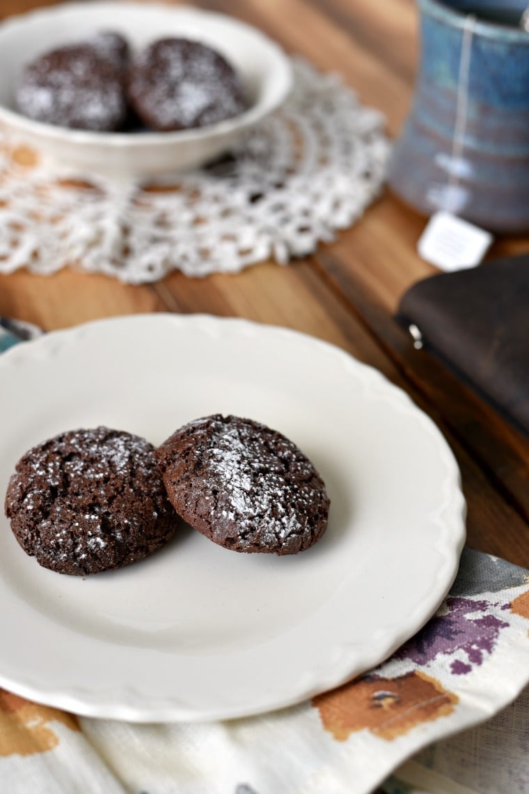 Two chocolate cookies dusted with powdered sugar sitting on a white plate on a wooden table with a book and ;ace doily next to it