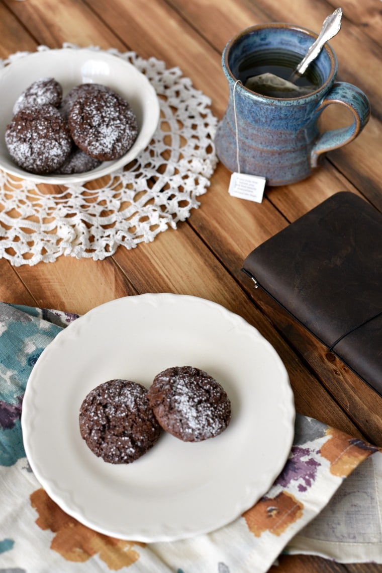 A white plate on a floral napkin, and a white bowl on a lace doily, both filled with chocolate cookies. There is a mug of tea and book beside