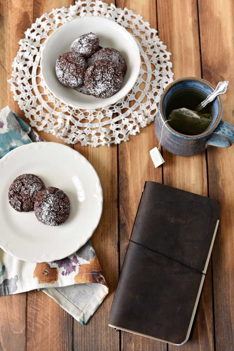 A wooden table with a plate and bowl of chocolate cookies on top. A mug of tea with a spoon and a leather bound book are also on the table