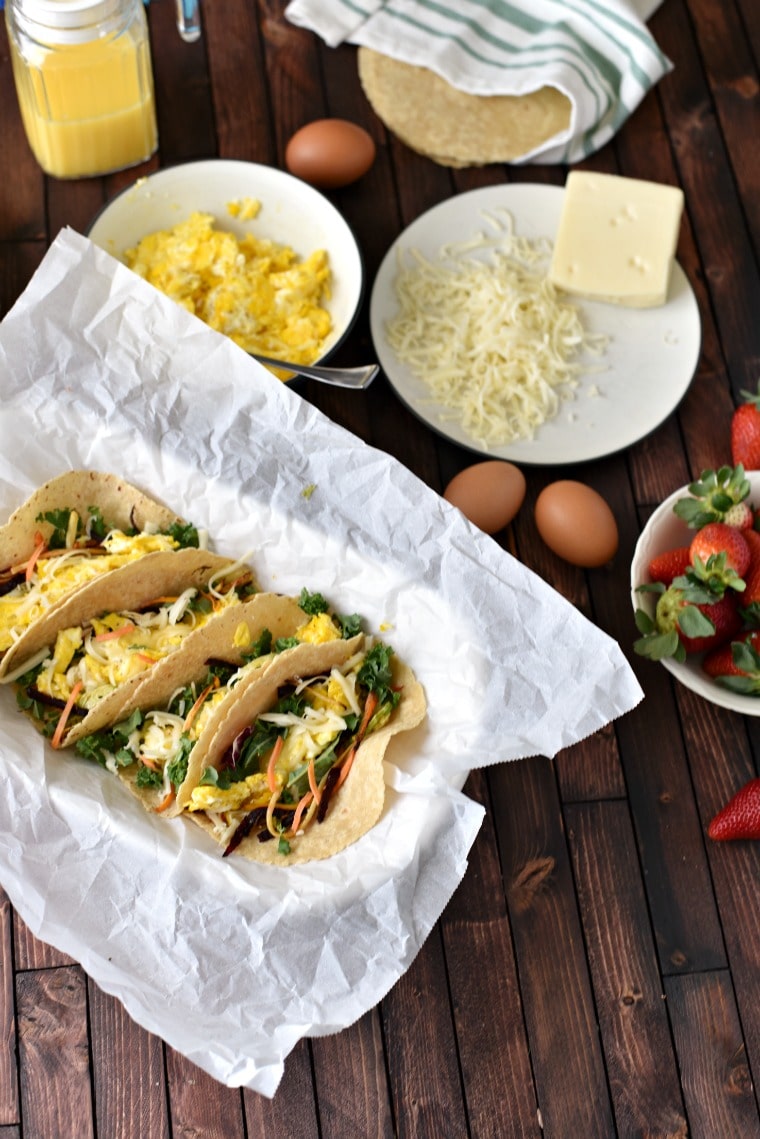 Four breakfast tacos on a plate covered in parchment paper on a wooden table. Beside are whole eggs, a bowl of strawberries, a bowl of scrambled eggs, & cheese