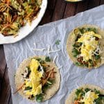 three breakfast tacos on a napkin on a wooden table next to a plate of shredded vegetables