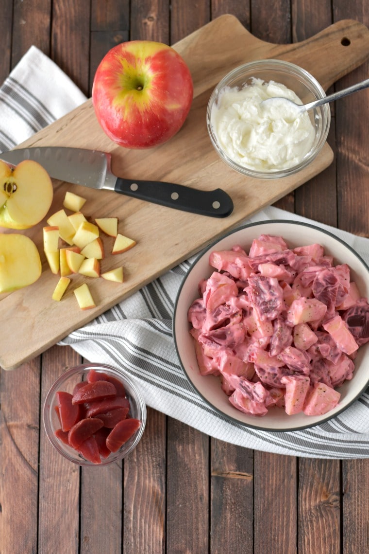 A wooden cutting board with a knife and chopped apples next to a bowl of beet and apple salad. All sitting on a black and white striped kitchen towel