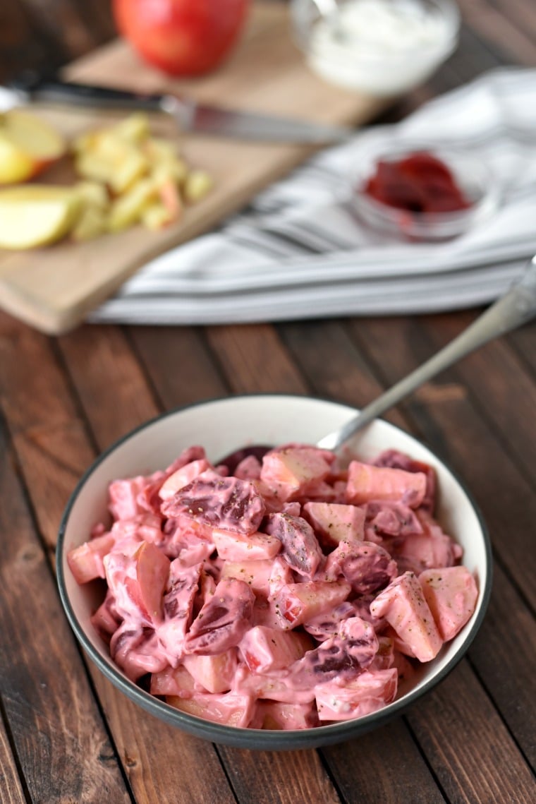 A spoon in a bowl of apple and beet salad with a cutting board with apple slices and a clear bowl of sliced beets blurred in the background