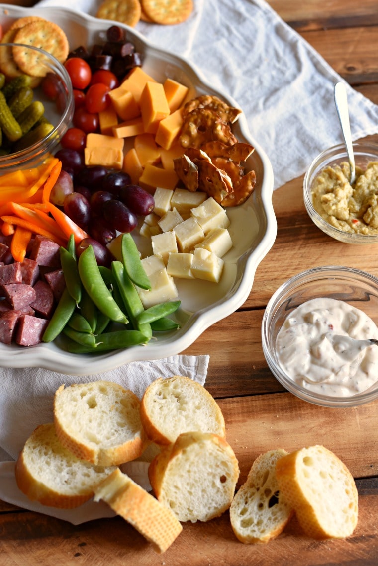 An assortment of snack foods on a white plate with sliced breads and two clear bowls of dips next to them