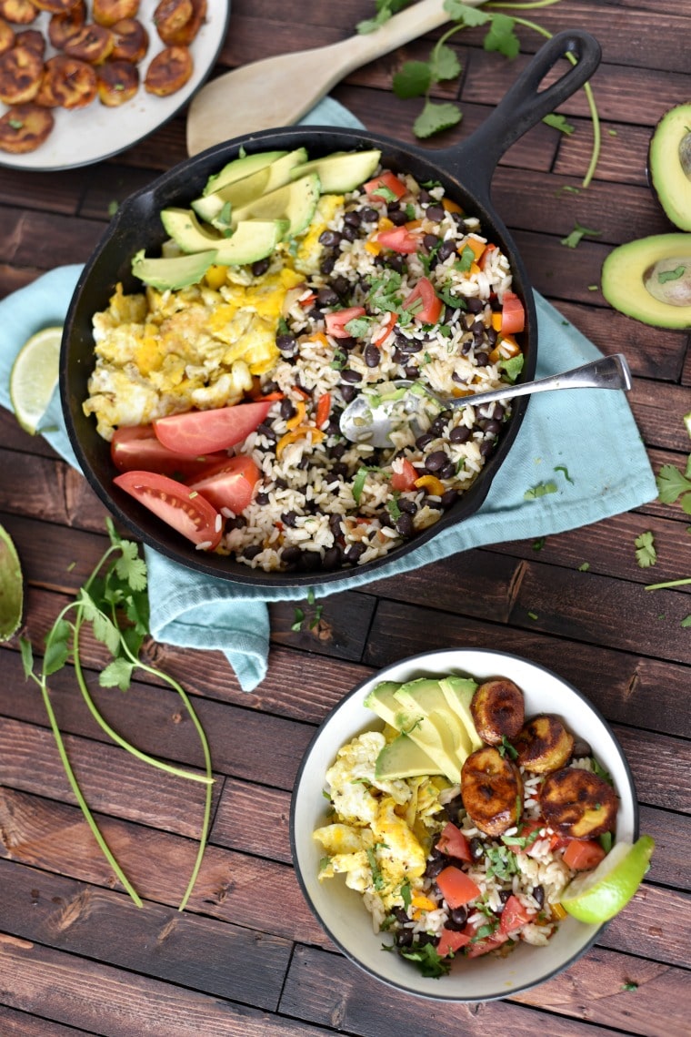 A cast iron skillet on a blue napkin and a white bowl on a wooden table. Both are filled with gallo pinto, a rice and beans food dish