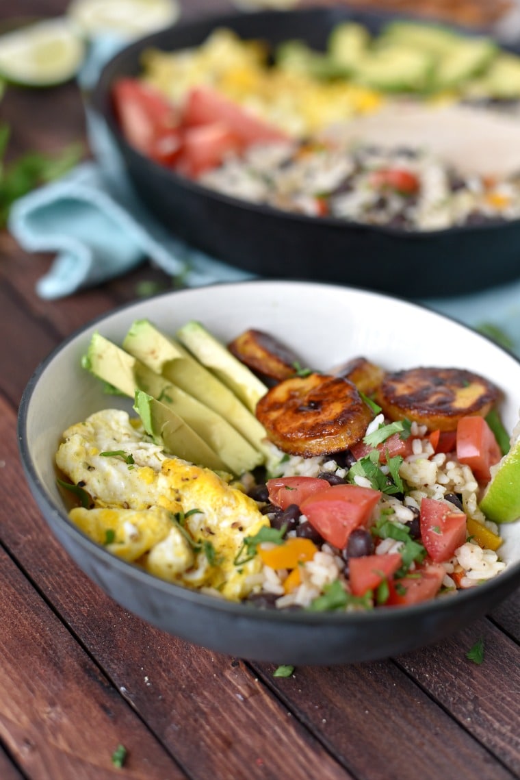 A bowl of rice with egg, tomato, black beans, sliced avocado, and fried plantains in the foreground, a cast iron skillet in the background