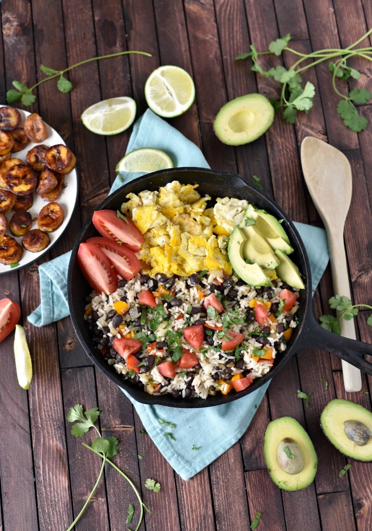 A cast iron skillet filled with gallo pinto, sitting on a blue napkin on a wooden table, surrounded by limes, halved avocado, and fried plantains