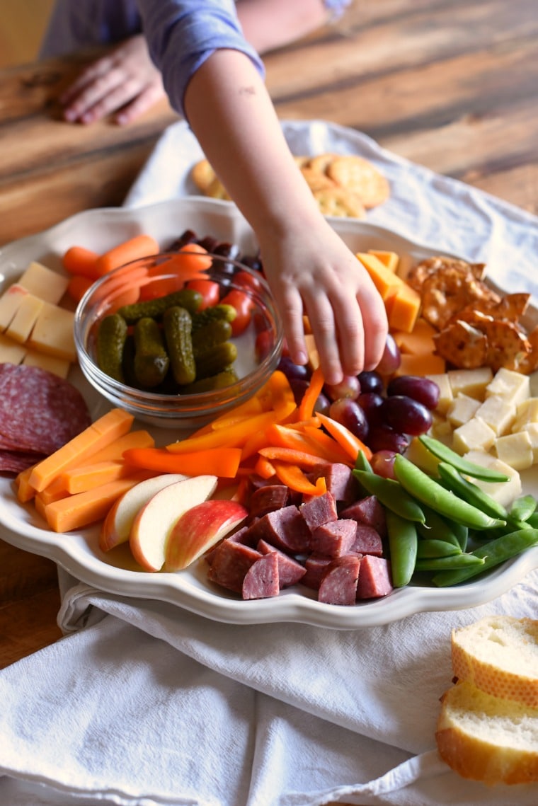 A small child reaching for grapes on a charcuterie plate filled with fruits, veggies, meats, and cheeses