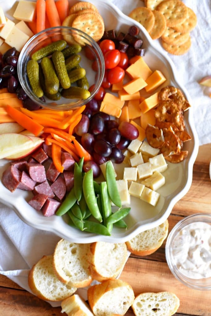 A white plate filled with cubed meats, cheeses, fruits, and veggies on a wooden table