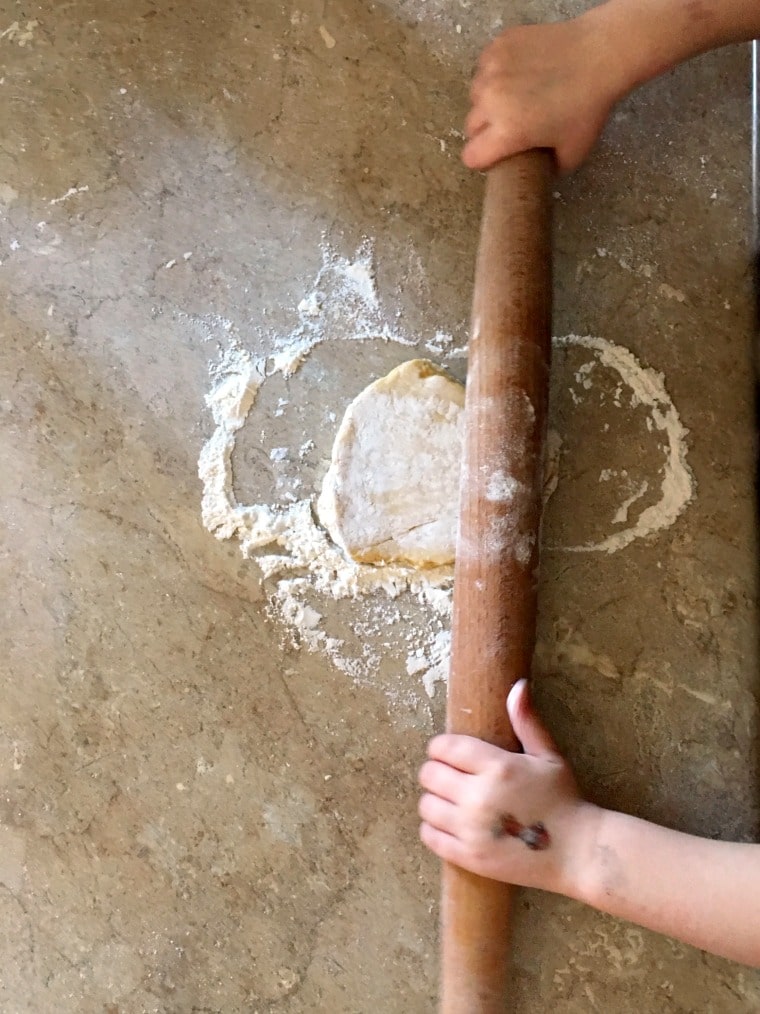 A child rolling out pasta dough on a tan countertop dusted in flour, using a brown wooden rolling pin