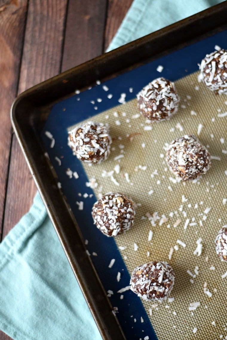 Chocolate coconut ball cookies on a baking sheet with a silicone baking mat. There is coconut sprinkled all around the baking mat. It is all on a wooden table