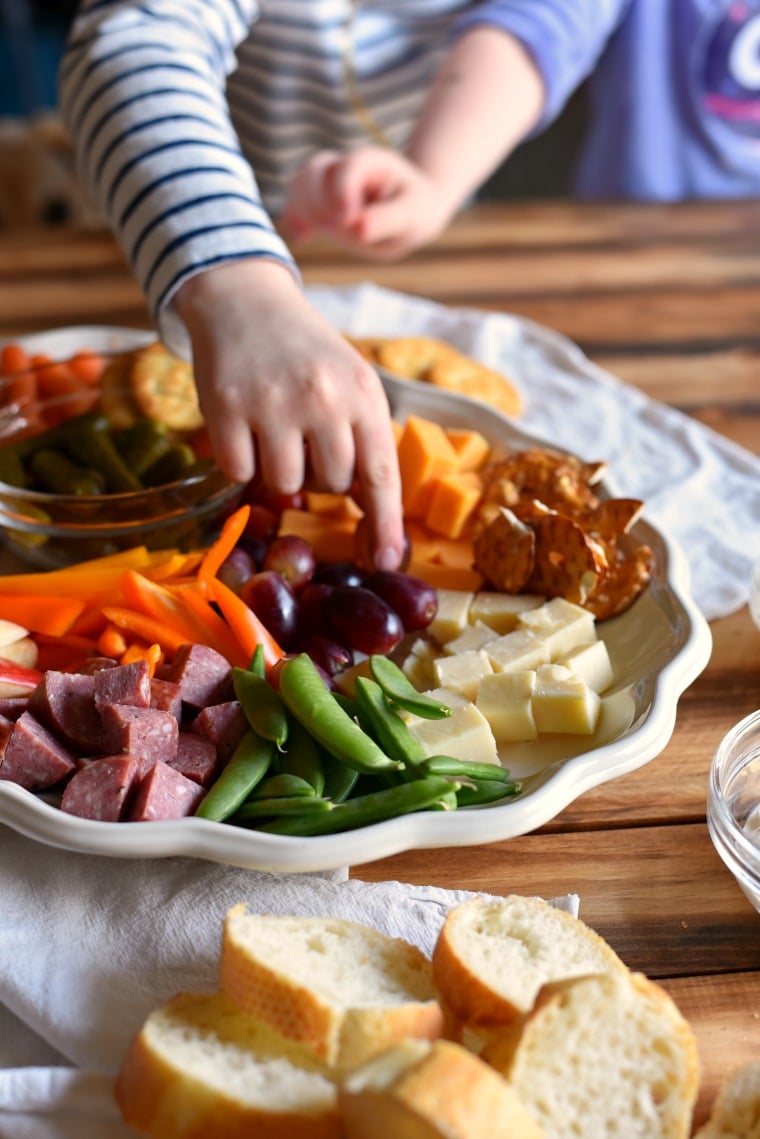 A small child in a striped shirt reaching for grapes on a plate filled with different kinds of meats, cheeses, fruits, and veggies