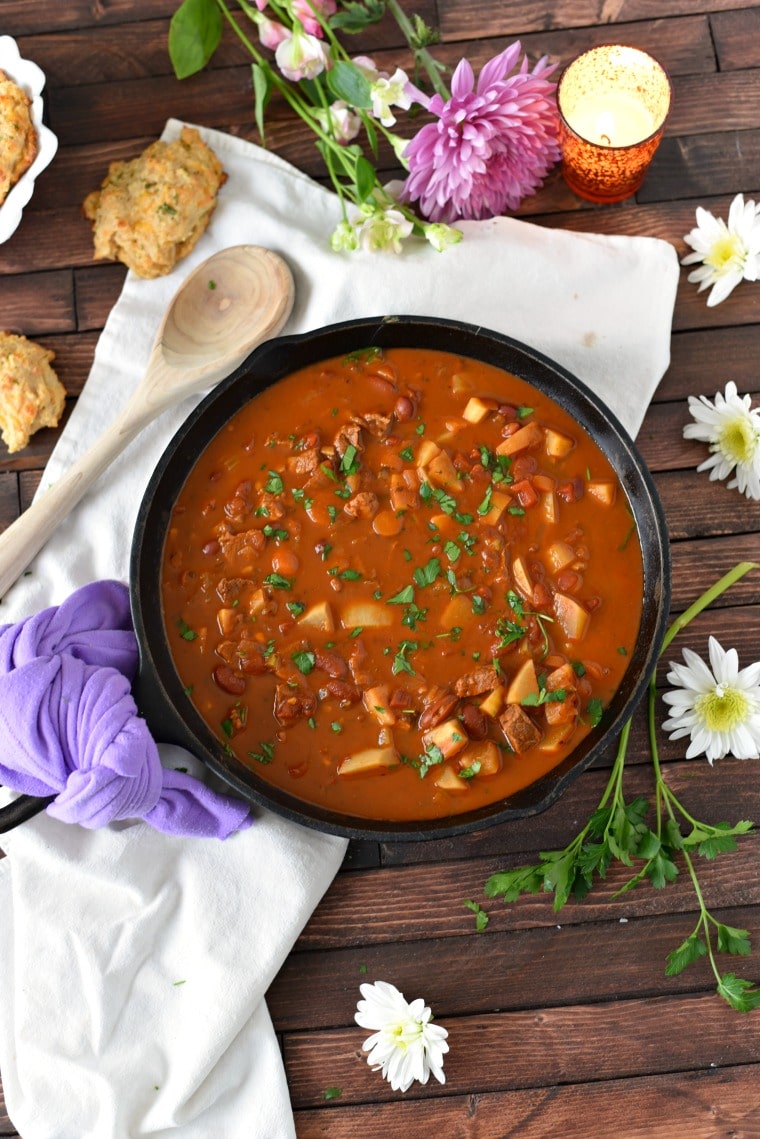 A bowl of goulash in a cast iron skillet sitting on a white napkin, surrounded by daisies and a wooden spoon