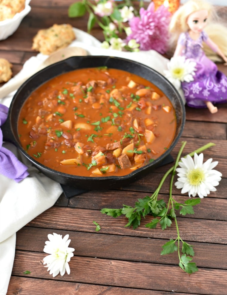 A cast iron skillet full of goulash with a Rapunzel doll in the background and daisies and herbs in the foreground