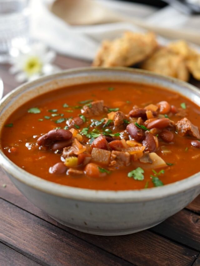 Goulash in a white bowl on a wooden table with biscuits and daisies in the background