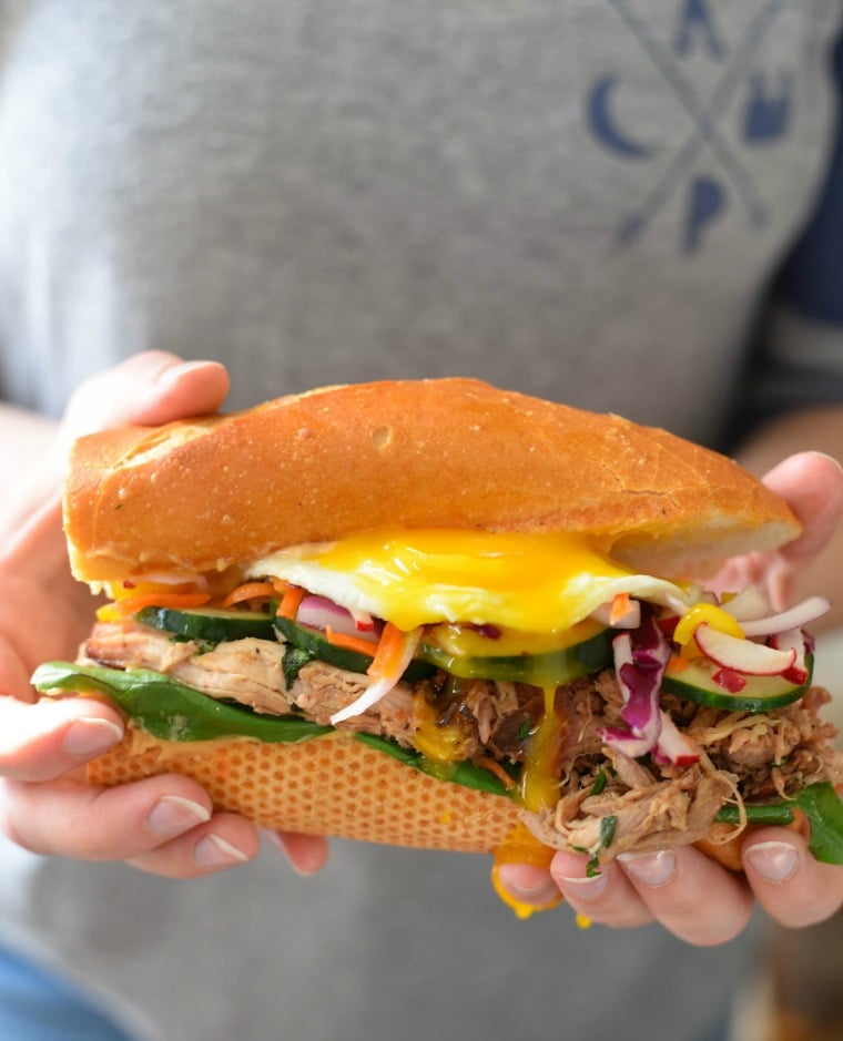 Hands holding a pork bahn mi sandwich with a runny egg