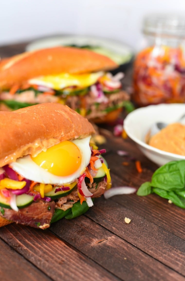 Two pulled pork bahn mi Sandwiches beside a boy of sriracha lime mayo both topped with eggs
