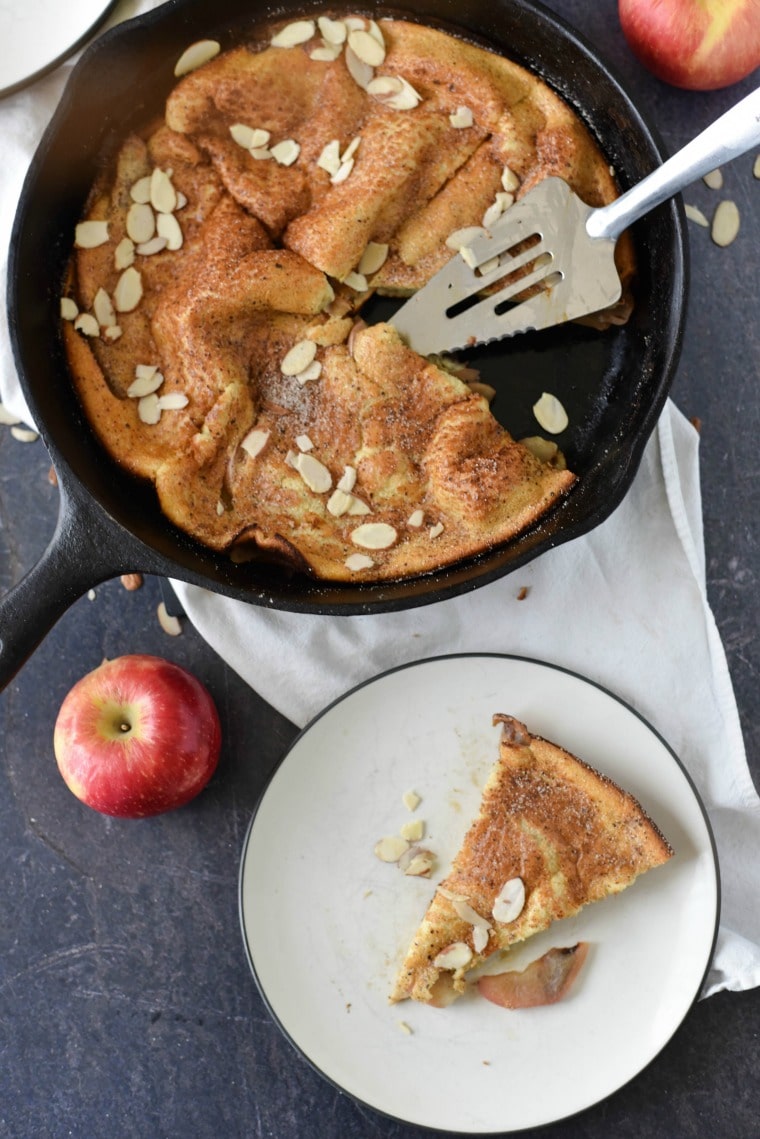 A slice of apple spiced dutch baby on a plate next to a cast iron skillet and apples 