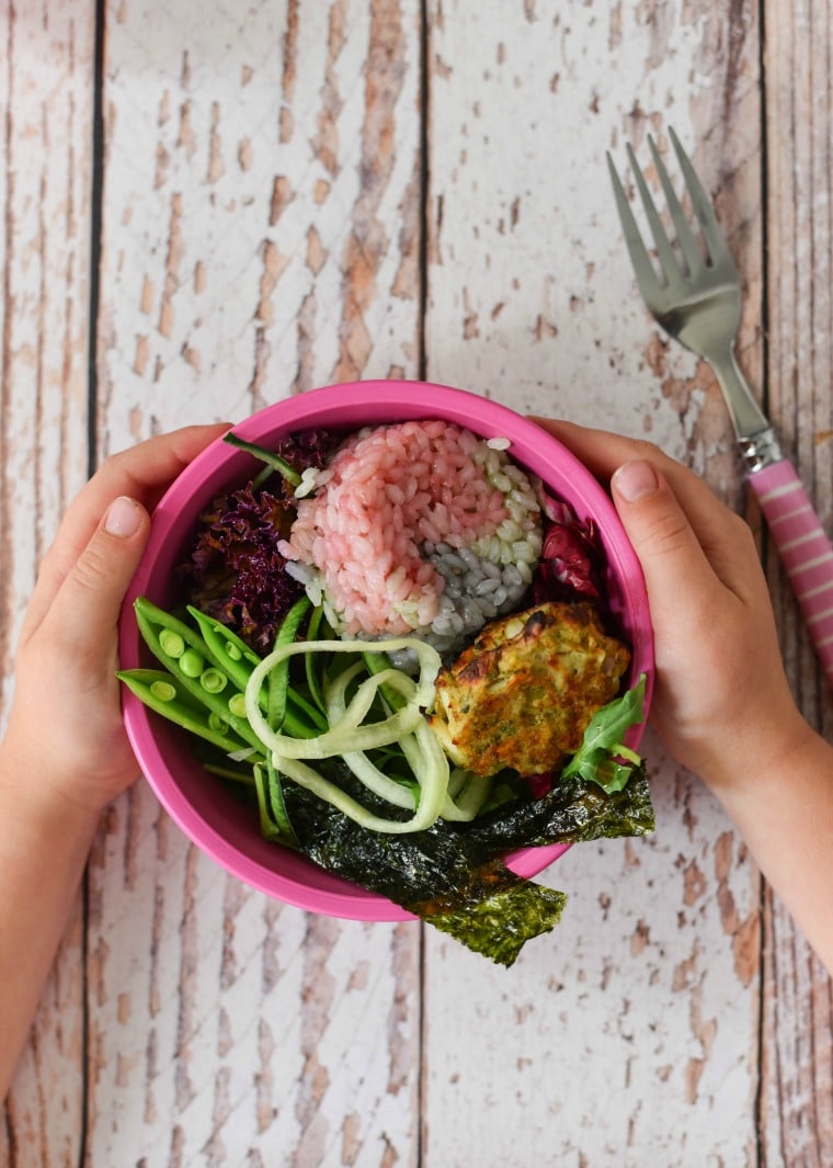 small pink bowl held in childs hands, filled with rainbow rice, vegetables and crab cake