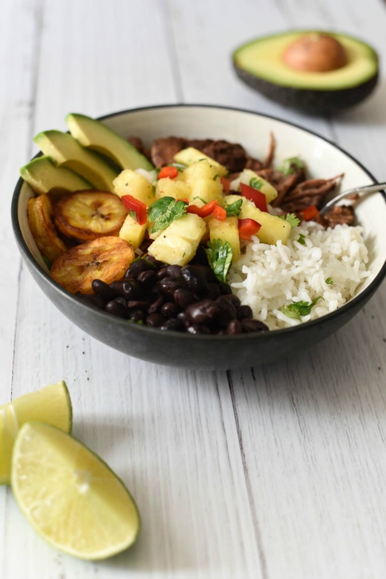 bowl with shredded beef, black beans, rice, fried plantains, avocado slices, and pineapple salsa next to lime slices
