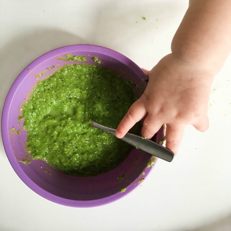 purple bowl filled with pureed green beans and baby hand reaching for spoon