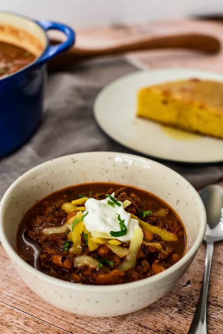 bowl of ground bison chili with chocolate with plate of cornbread and pot of more chili behind