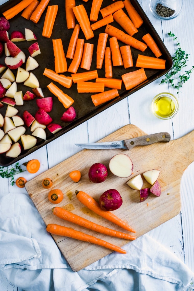 A wooden cutting board with carrots and potatoes next to a sheet pan