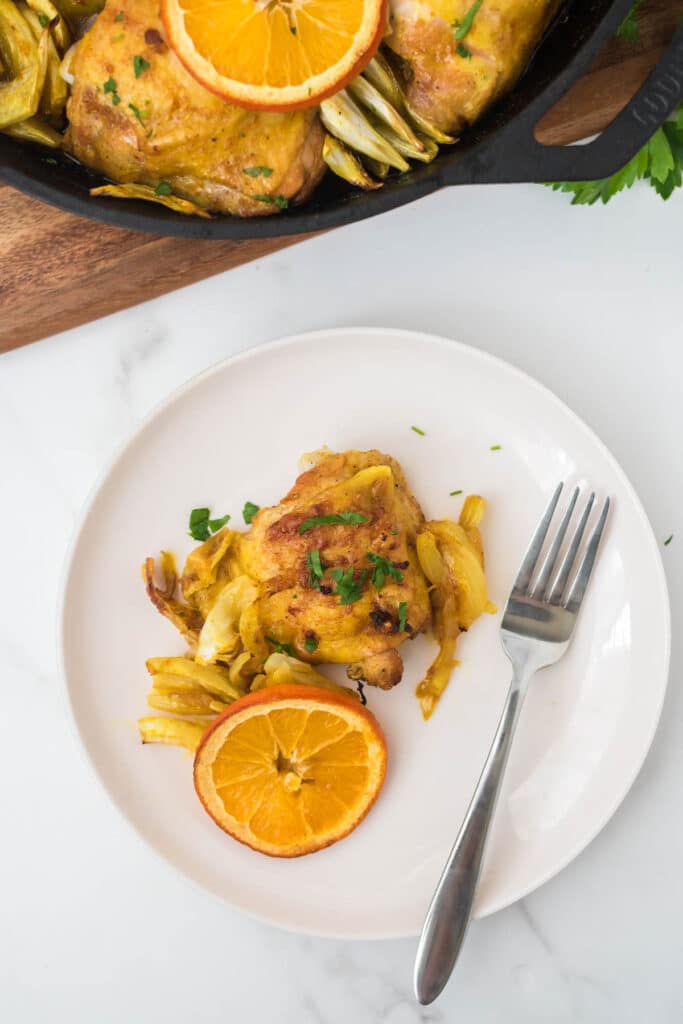 white plate with turmeric chicken thigh and orange slice on it with a fork, with cast iron skillet with more at top of screen