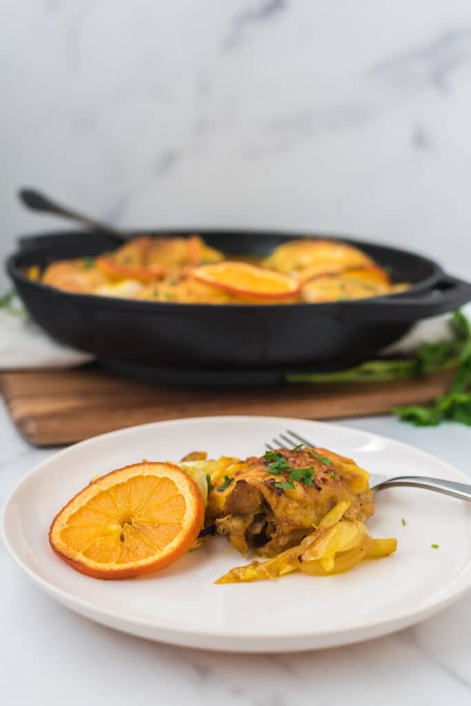 plate of roasted chicken thighs with fennel and orange slices in front of cast iron skillet with more chicken and serving spoon
