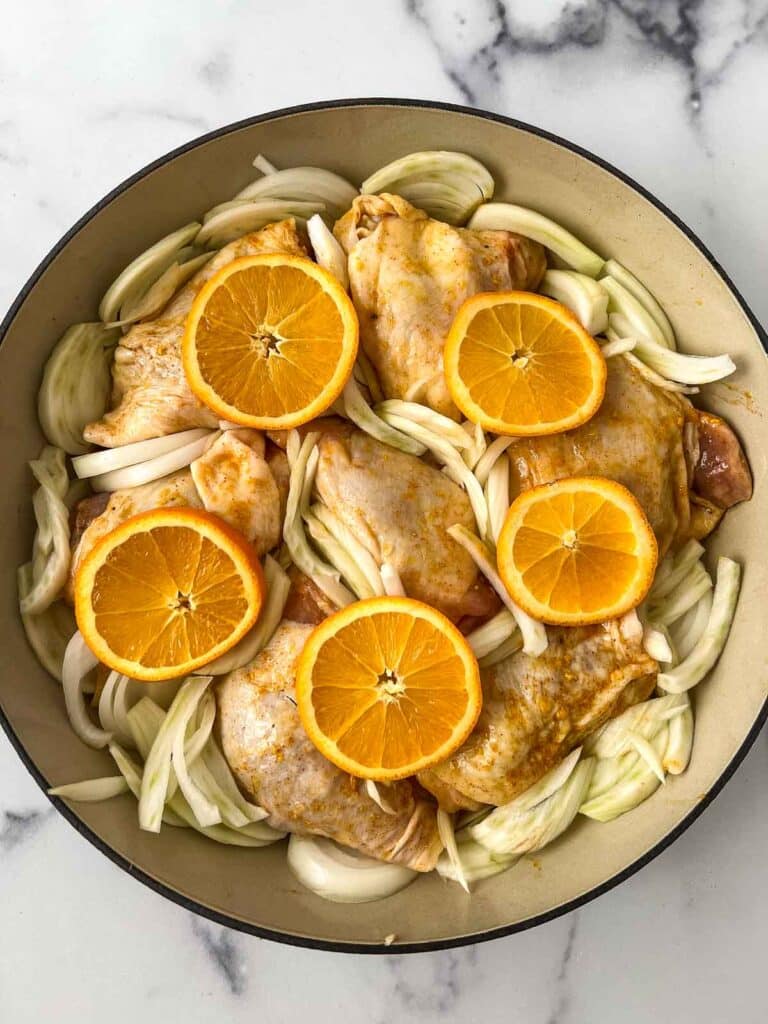 circle baking pan with chicken thighs, onion, fennel slices, and orange slices with marinade