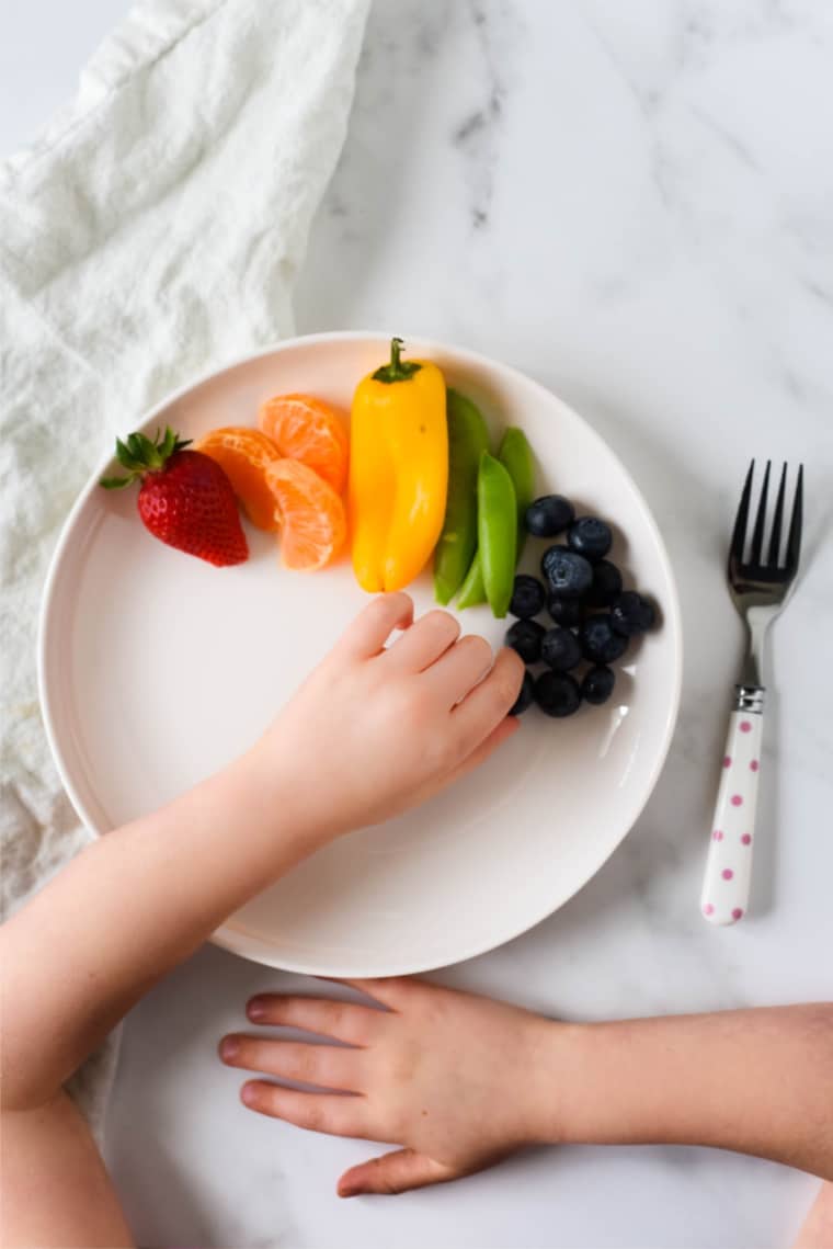white plate with rainbow produce and kids hands sneaking bite