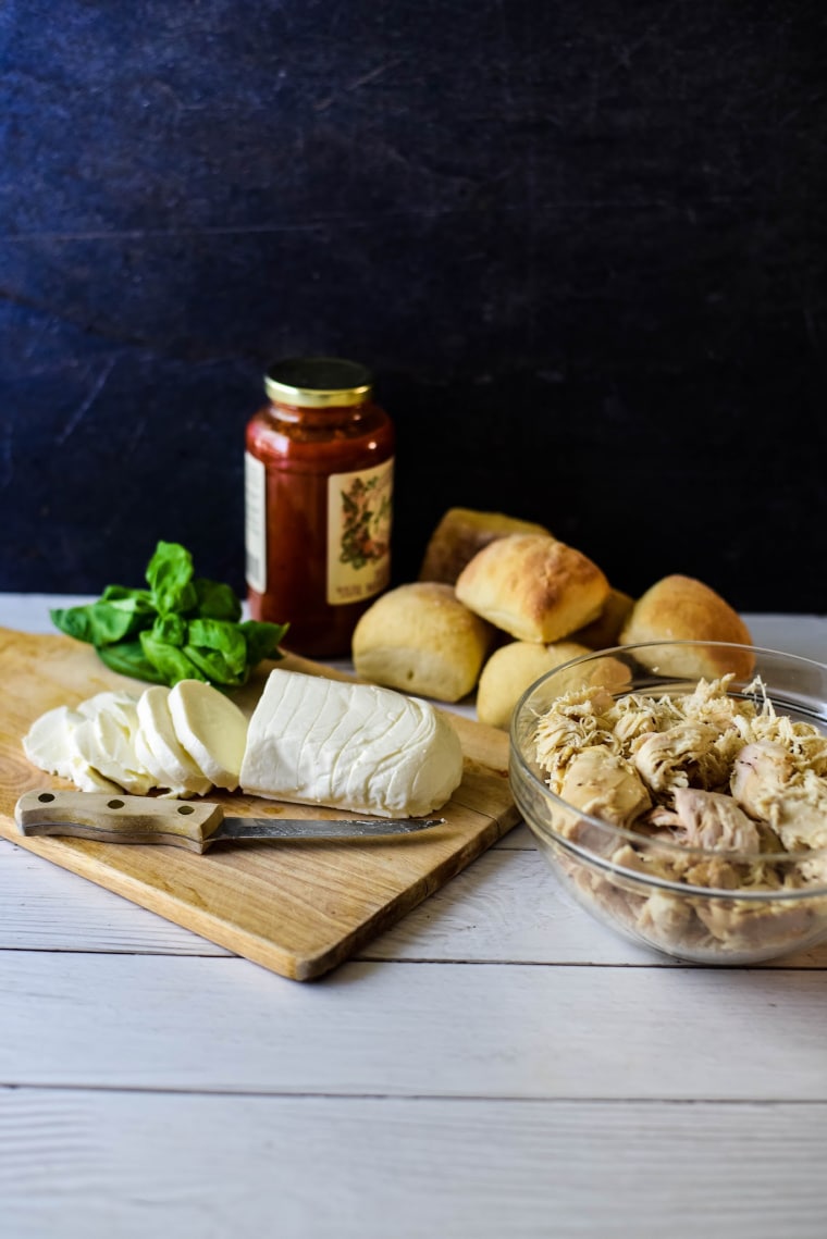 wooden cutting board with bowl of shredded chicken, sliced mozzarella, fresh basil, cabana rolls and a jar of marinara sauce arranged around