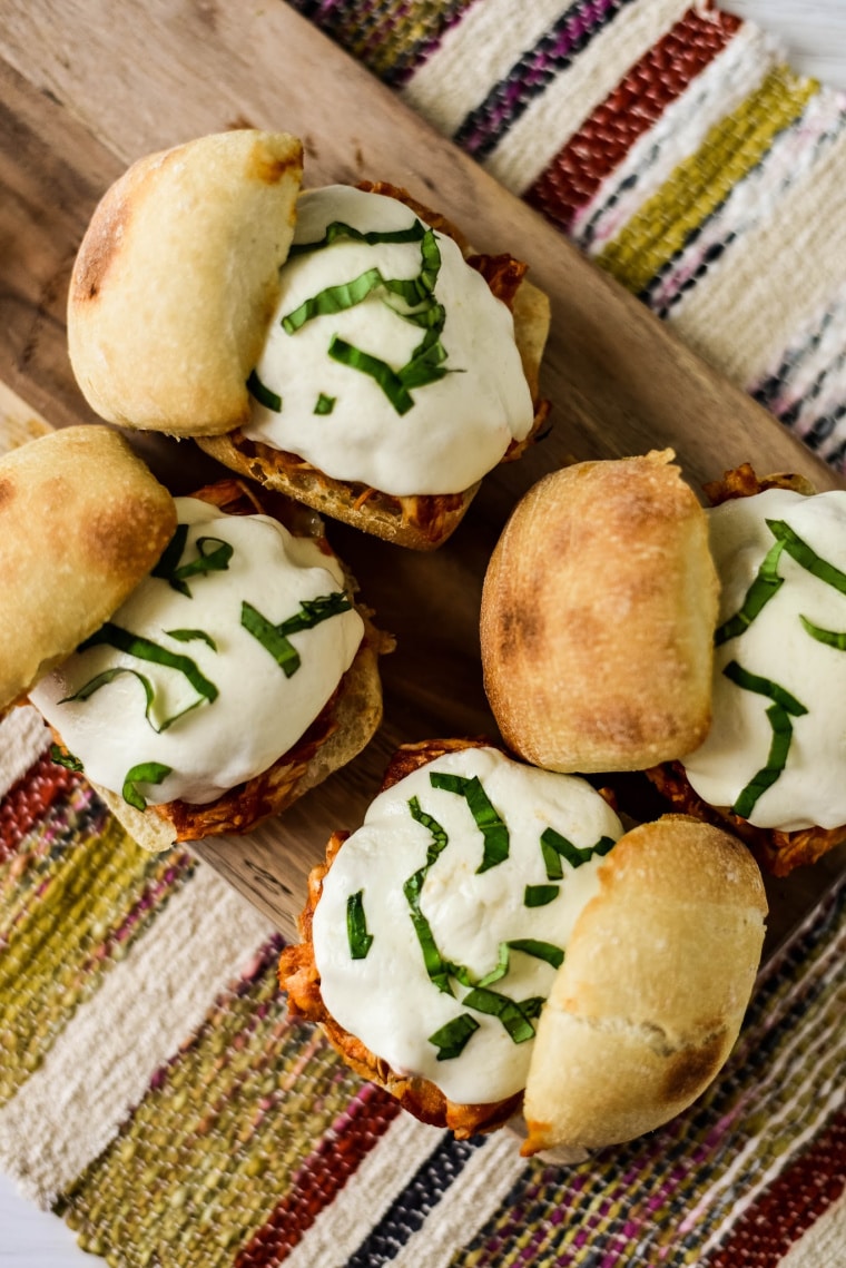 four chicken parmesan sandwiches on wooden board with tops askew, showing melty cheese and sliced basil