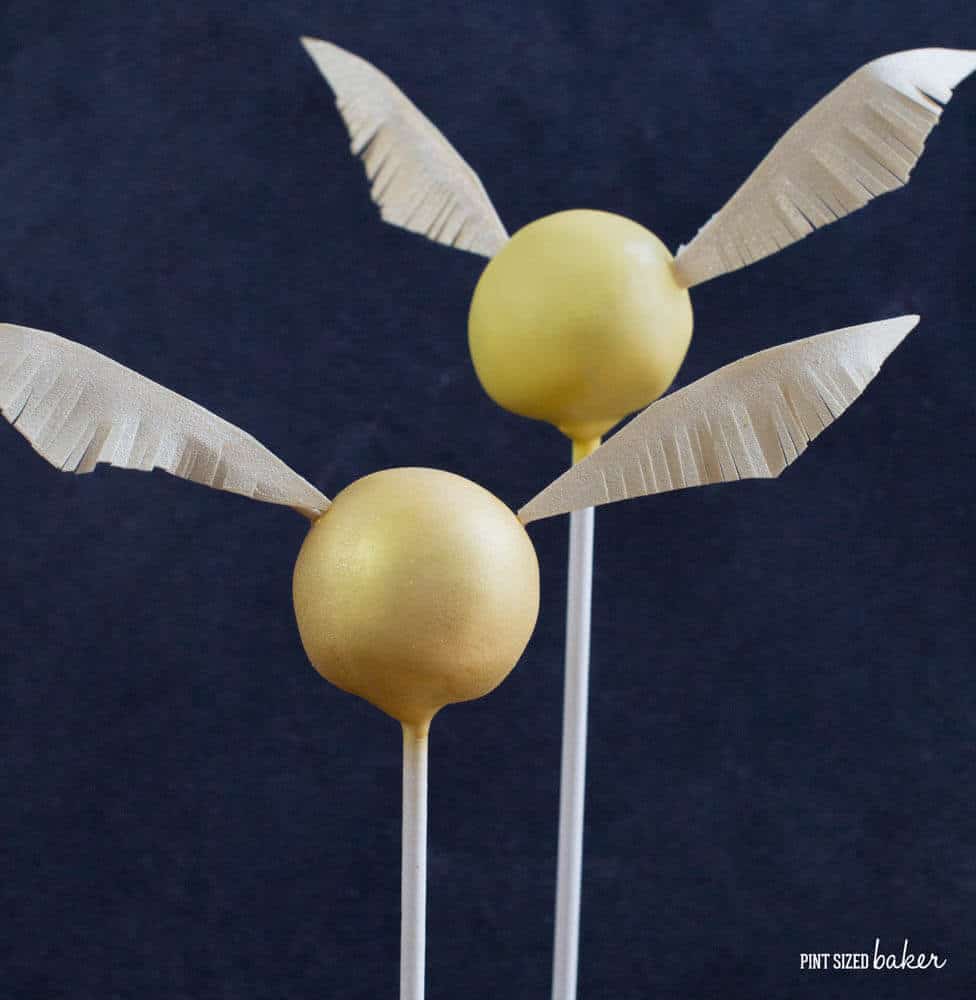 two cake balls on sticks with golden frosting and wings made to look like a snitch from Harry Potter books