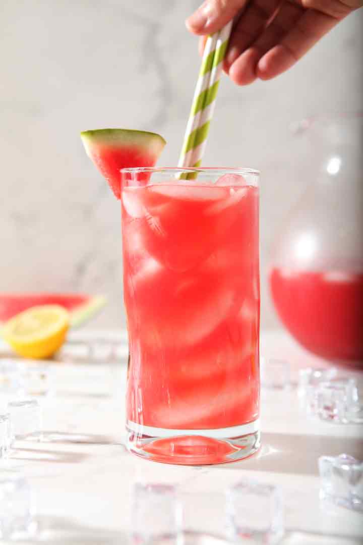 agua fresca in tall glass with slice of watermelon on rim of glass and hand stirring straw