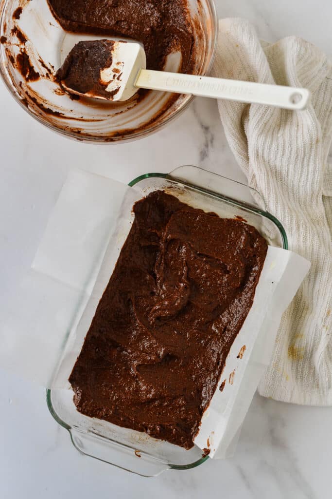 glass baking dish with brownie batter next to empty dirty bowl and rubber spatula