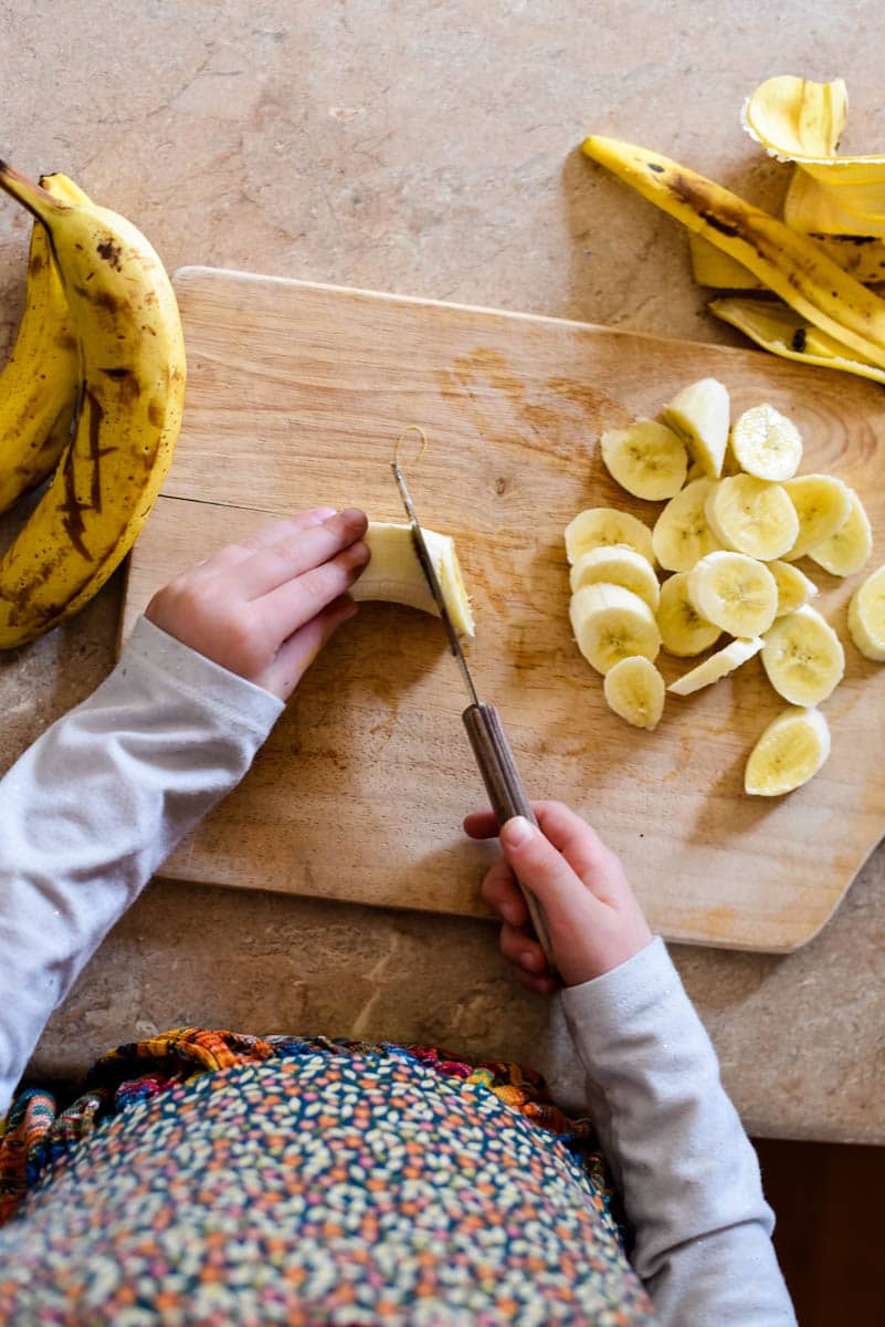 childs hands slicing bananas on wooden cutting board