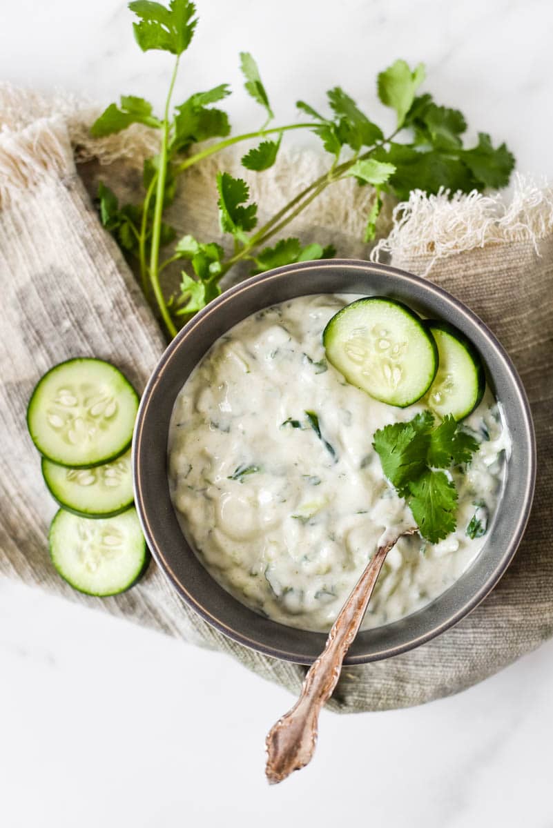 grey bowl filled with cucumber raita with sliced cucumbers and cilantro beside.