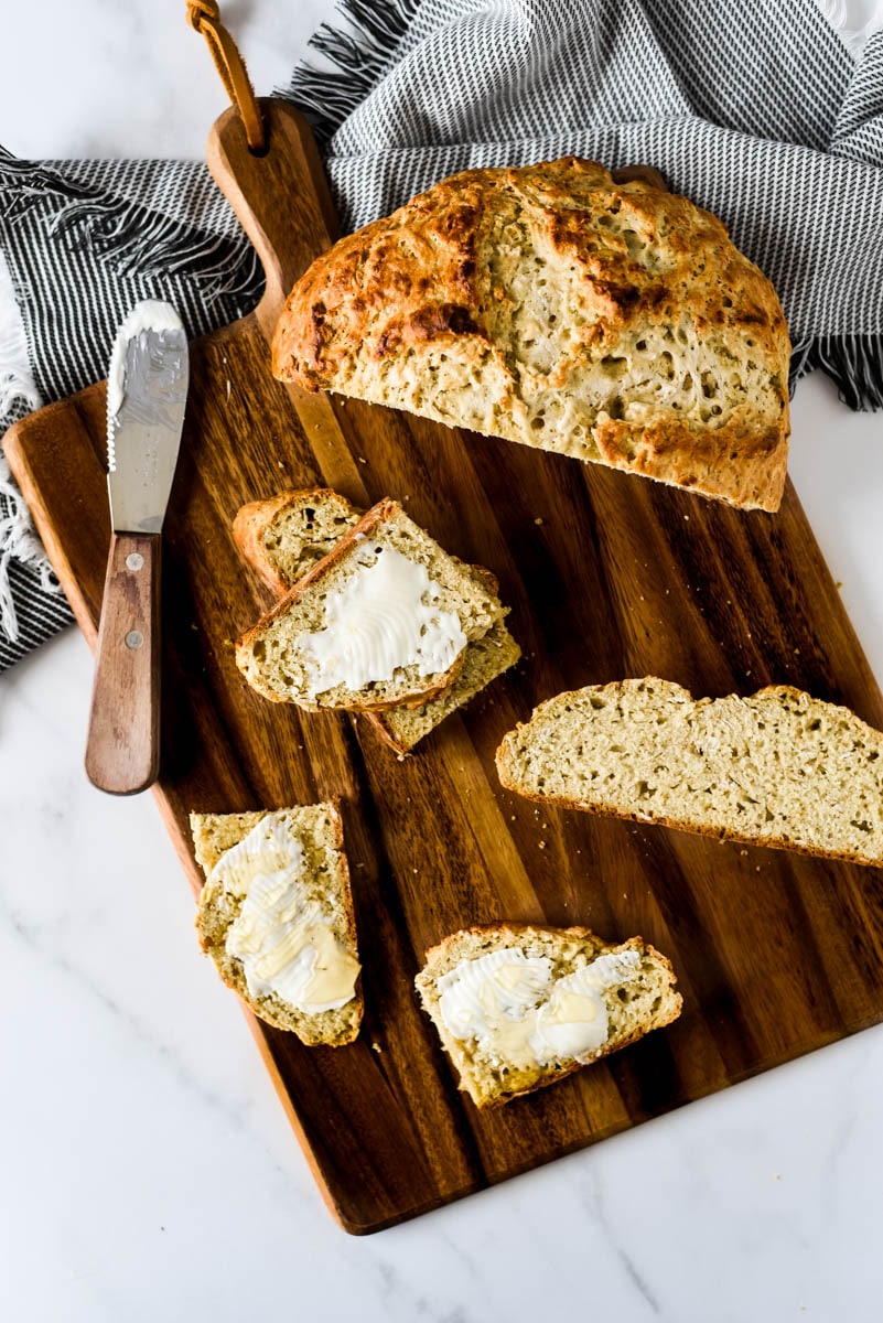 wooden cutting board with bread knife, on it is a loaf of sourdough soda bread with some slices cut and buttered and drizzled in honey