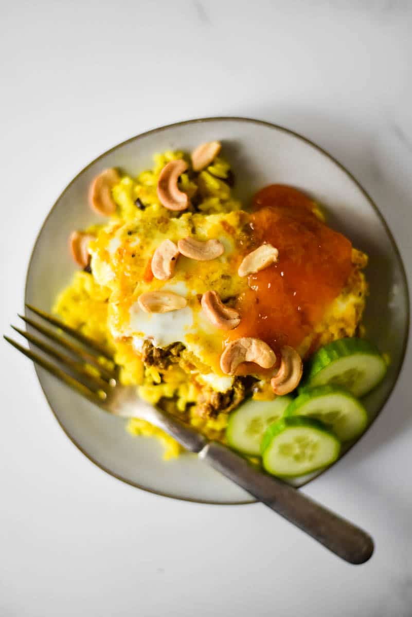 grey plate with bobotie, yellow rice, sliced cucumbers, cashews and apricot jam. Fork on the side of the plate.