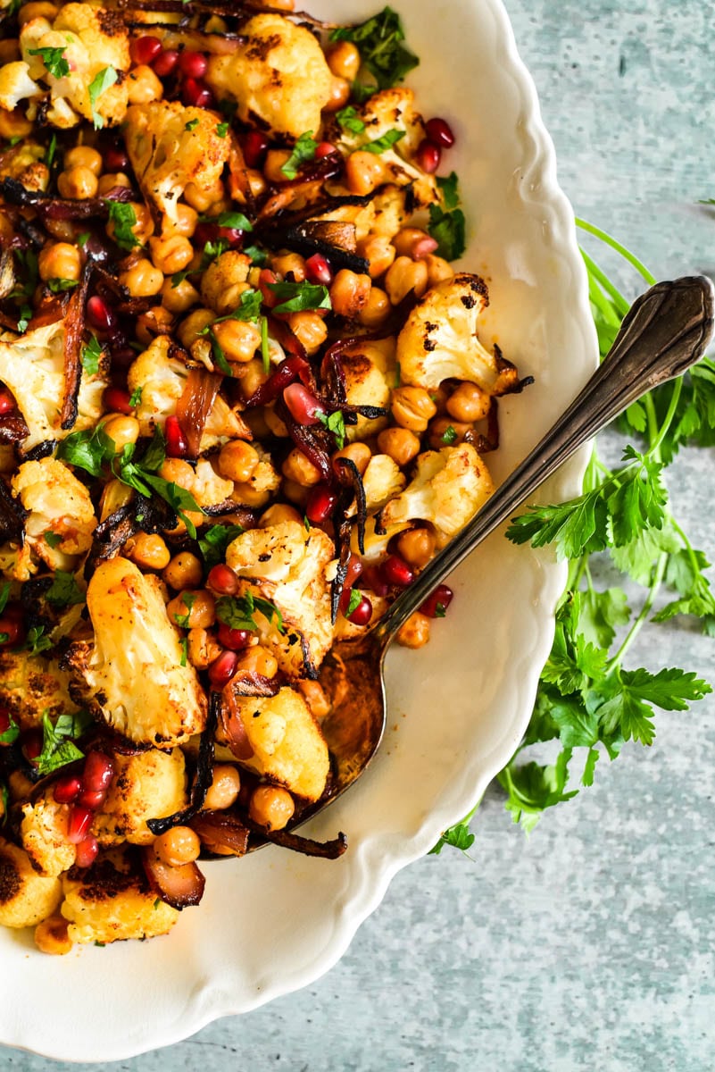 close up overhead view of roasted cauliflower with chickpeas onions and pomegranates  on white platter with serving spoon to the side