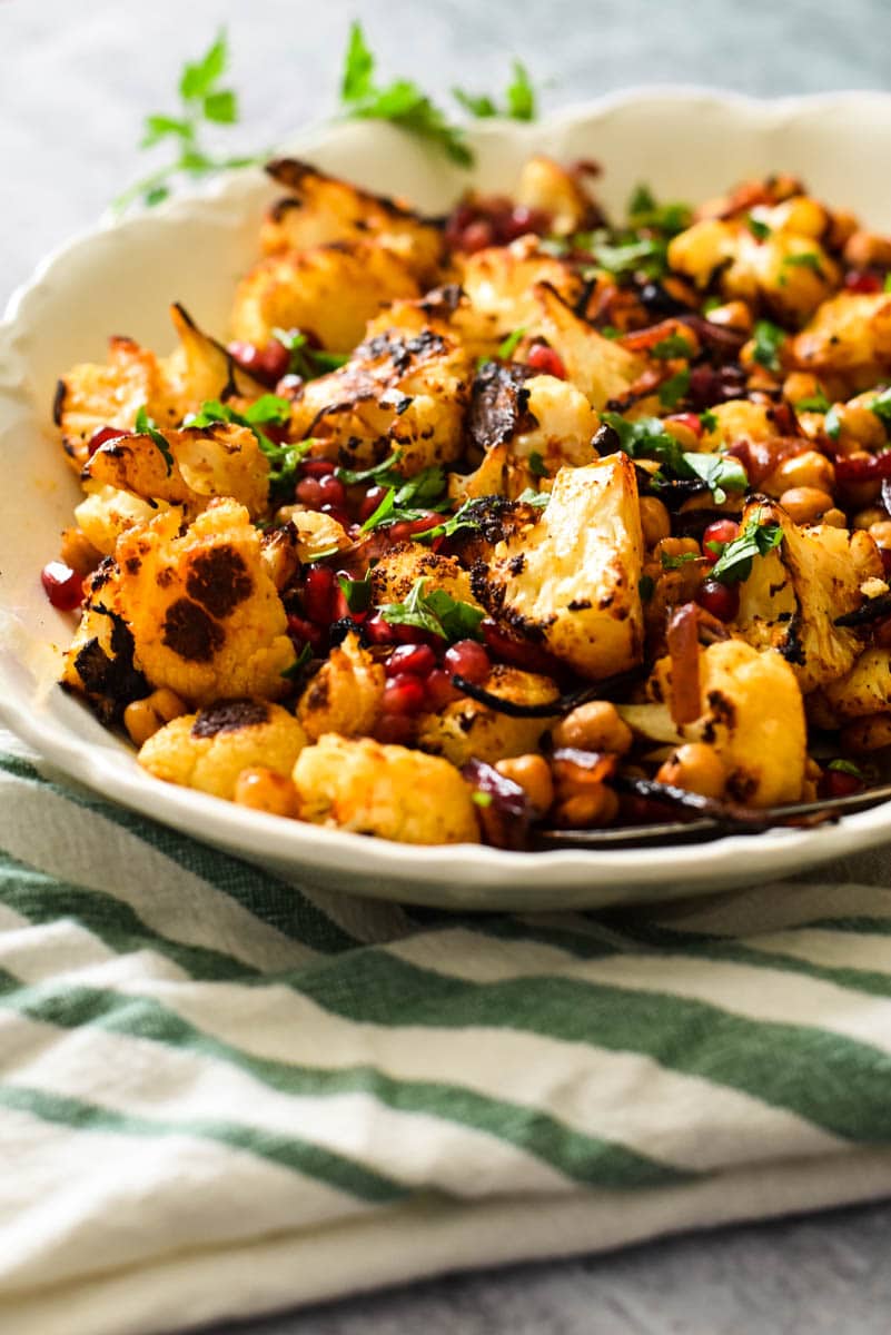 side view of white platter with roasted cauliflower and onions topped with pomegranates and parsley, with striped kitchen towel next to platter