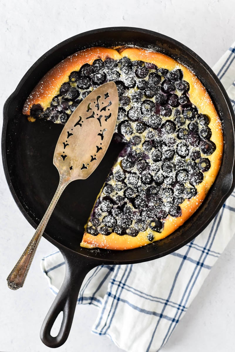 cast iron skillet with blueberry clafoutis with several pieces missing and serving spatula on top