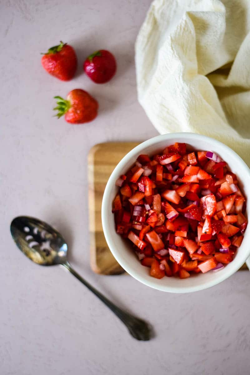 overhead of bowl of strawberry salsa with serving spoon and three strawberries on the side