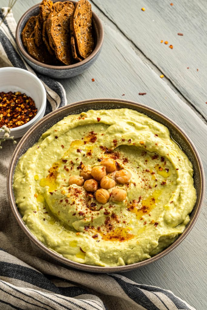 avocado hummus in bowl with crackers and spices behind