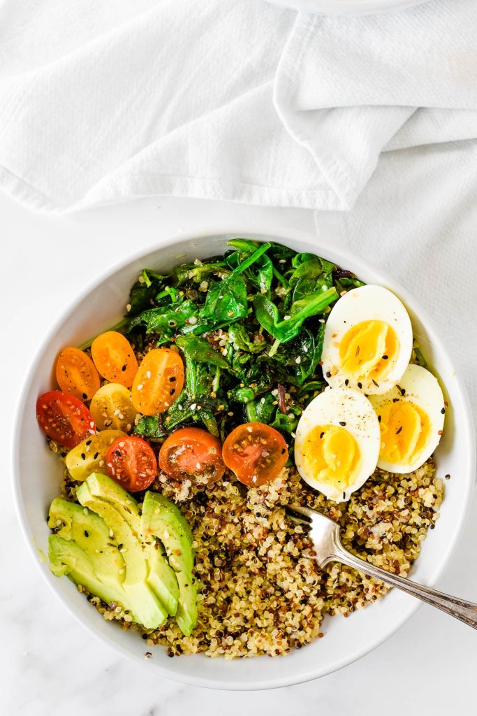 overhead image of white bowl with quinoa, hard boiled eggs, avocado slices, cherry tomatoes and sautéed kale