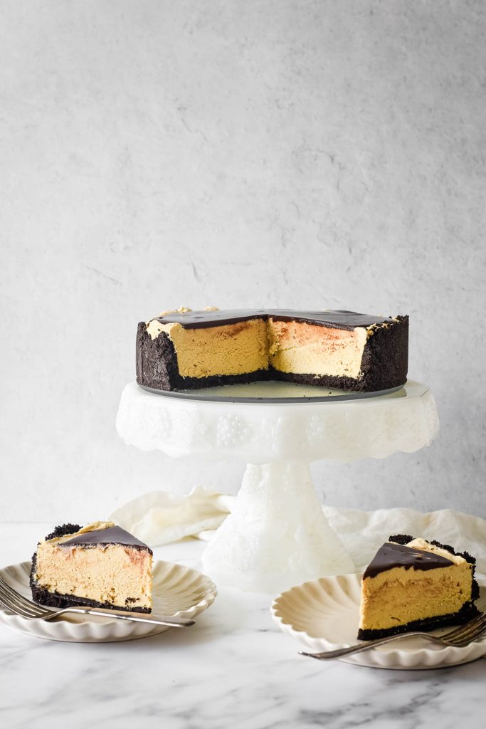 peanut butter cheesecake on white cake stand with two slices on plates besides