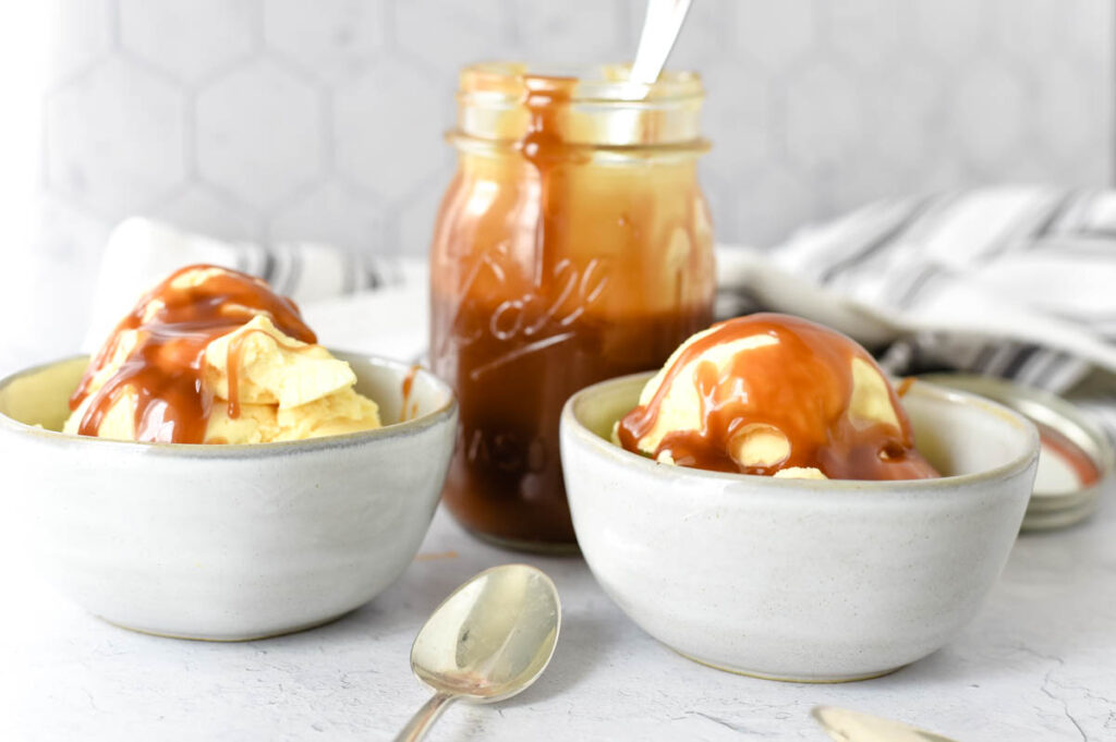 two bowls of ice cream with caramel sauce on top and jar of sauce behind