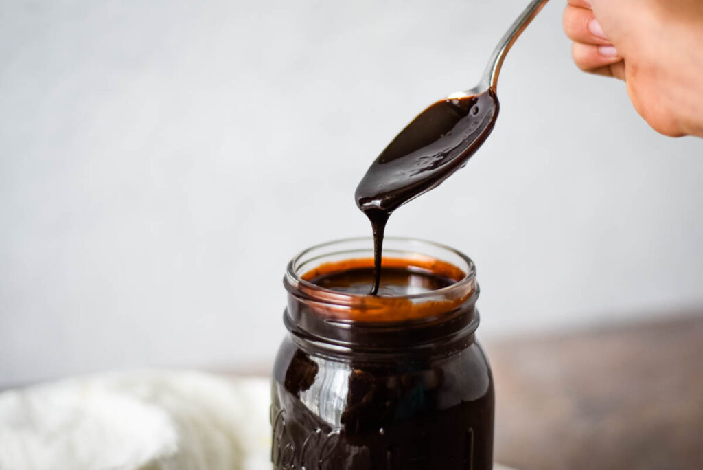 child's hand holding spoon over mason jar of hot fudge sauce, sauce drizzling off spoon and back into jar