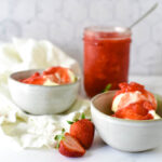 two bowls of ice cream topped with strawberry sauce and jar of sauce in background